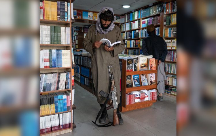 A Taliban fighter stands in a library, reading a book with his AK-47 between his legs, reflecting the group's control over educational content in Afghanistan. (Photo Credit: Social Media)