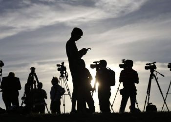 Journalists capture pivotal moments under the looming threat of the Taliban, their identities shrouded in anonymity against the backdrop of a vast sky and faint sunlight.