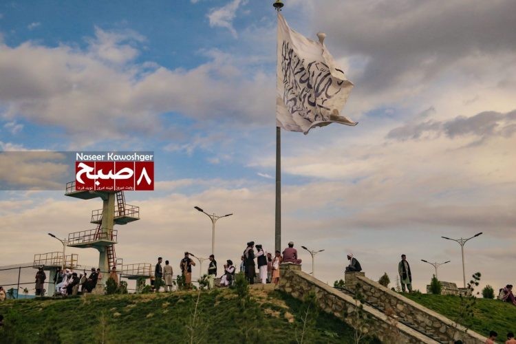 Large Taliban flag displayed in a Kabul park, with groups of people roaming around, capturing daily life under Taliban governance. (Photo Credit: 8am.media)