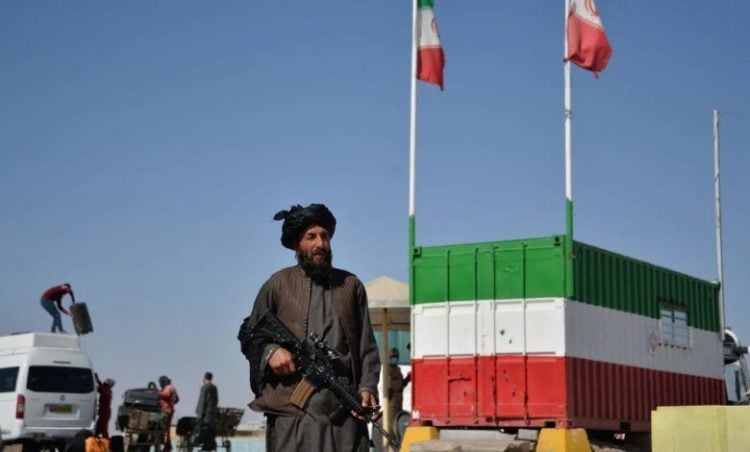 A Taliban militant stands guard along the Nimruz-Zahedan border as Iranian flags flutter on a nearby truck. Drivers on this route face economic hardship due to a recent suspension of cross-border transportation.