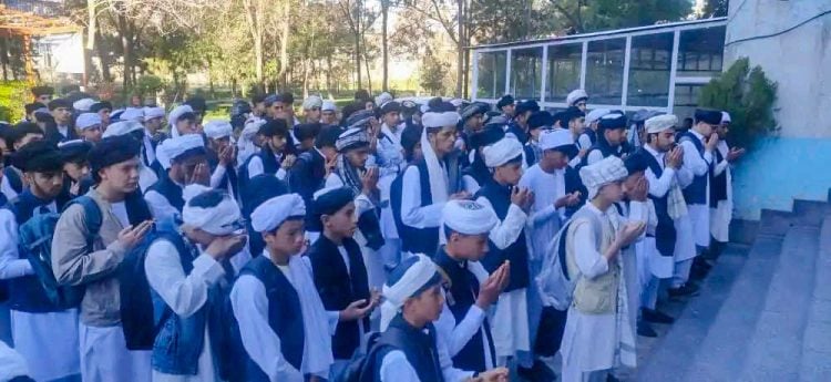 Taliban students in mandatory uniforms, including a white shirt, trousers, and headscarf (lungi), participating in a morning prayer at a school in Afghanistan.