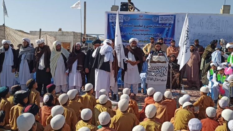 Taliban officials oversee a religious graduation ceremony at a madrasa in Afghanistan, with children in traditional attire seated before them under the group's flags.
