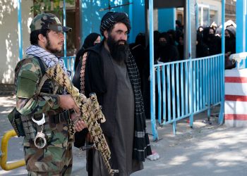 A Taliban fighter stands guard alongside a senior official as women, dressed in full coverings, wait behind a barrier outside a government building in Afghanistan, June 2025. (Photo Credit: AFP)
