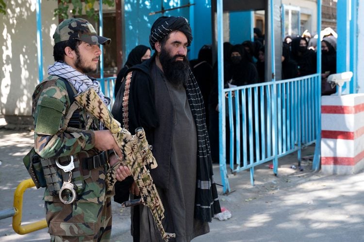 A Taliban fighter stands guard alongside a senior official as women, dressed in full coverings, wait behind a barrier outside a government building in Afghanistan, June 2025. (Photo Credit: AFP)