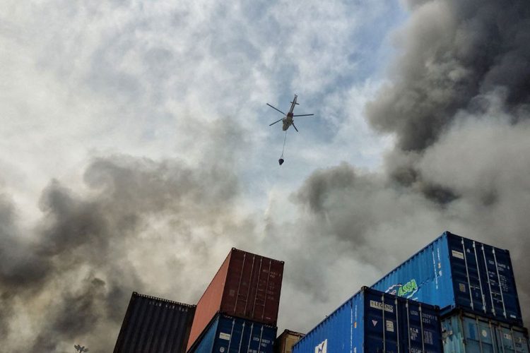 A helicopter hovers over shipping containers amidst thick smoke at Bandar Abbas port, highlighting the impact of recent conflicts on trade operations. (Photo Credit: AFP)