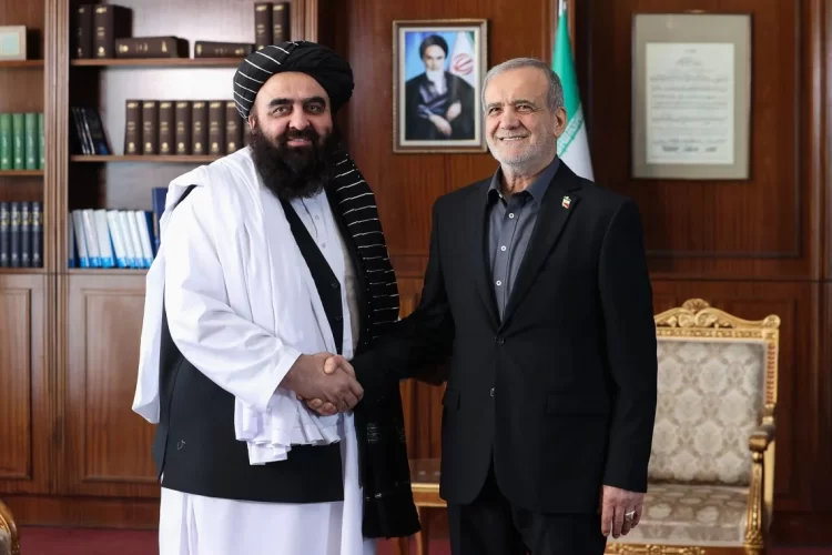 Mawlawi Amir Khan Muttaqi and Pezeshkian shake hands in a diplomatic meeting, with an Iranian flag and portrait in the background. (Photo Credit: AFP)
