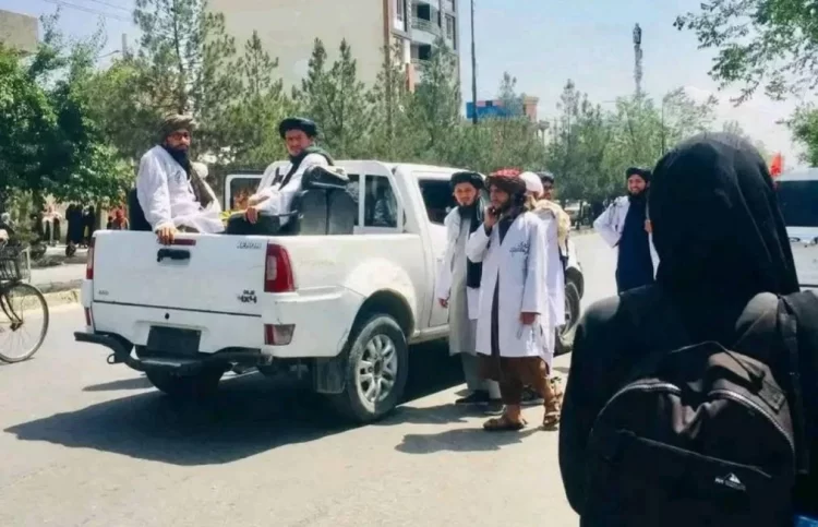 Taliban Morality Police patrol a street in Kabul, with armed individuals in a pickup truck and others standing nearby, as a woman with a backpack walks past. (Photo Credit: Social Media)