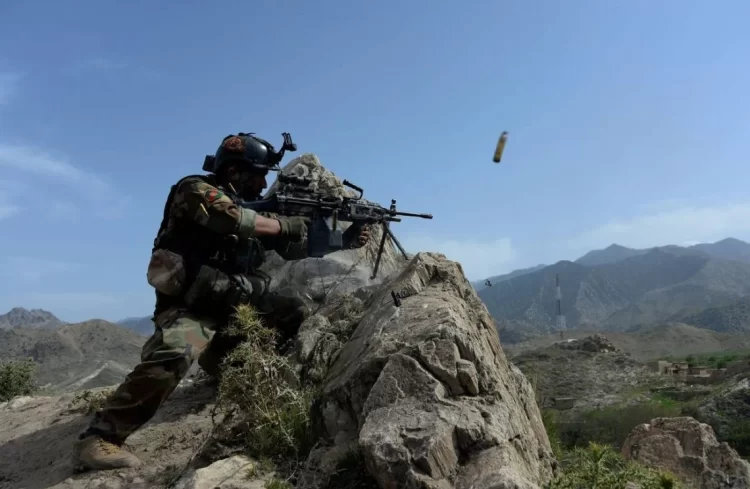 An Afghan soldier takes cover behind a rock, firing a machine gun in a rugged mountainous region, with a spent shell casing in mid-air. (Photo Credit: WSJ)