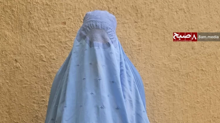 A woman in Afghanistan stands against a wall, wearing a blue burqa, symbolizing the restrictive decrees imposed under Taliban rule. (Photo Credit: 8am.media)