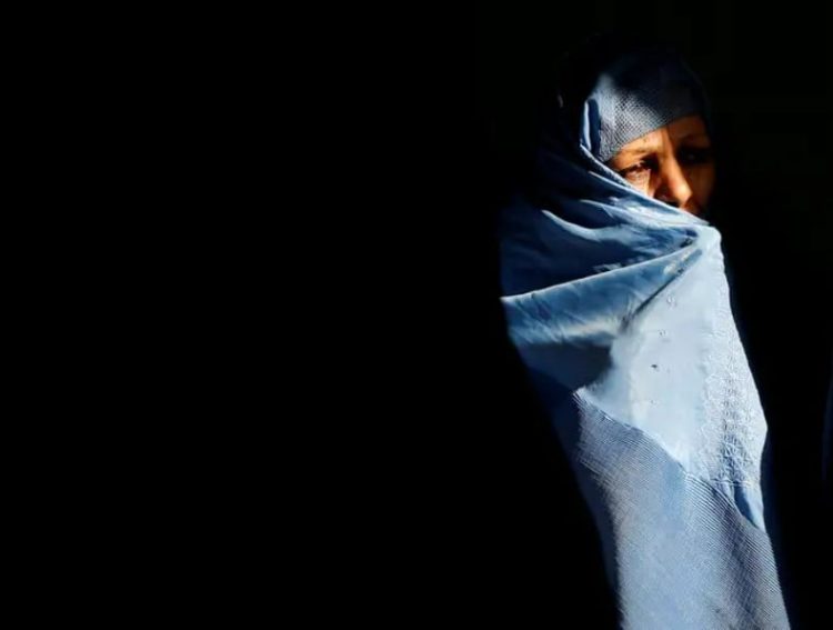 An Afghan woman in traditional blue burqa covering her face against a dark background, representing the severe restrictions faced by women in Taliban-controlled Afghanistan where property rights and inheritance are systematically denied. (Photo Credit: Reuters)