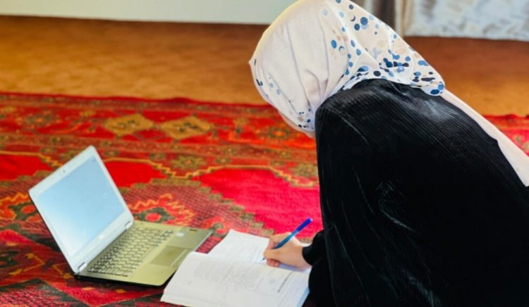 A young girl studies at home in Afghanistan using her laptop and notebook, representing the critical role of online education for girls who have been barred from attending school in person for over a thousand days. (Photo Credit: Embrace Relief)
