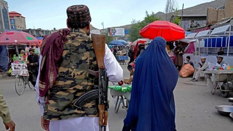 A woman in a blue burqa walks through a Kabul marketplace as a Taliban fighter in camouflage uniform stands nearby, illustrating the restrictive environment Afghan women navigate daily under Taliban rule. (Photo Credit: AP)