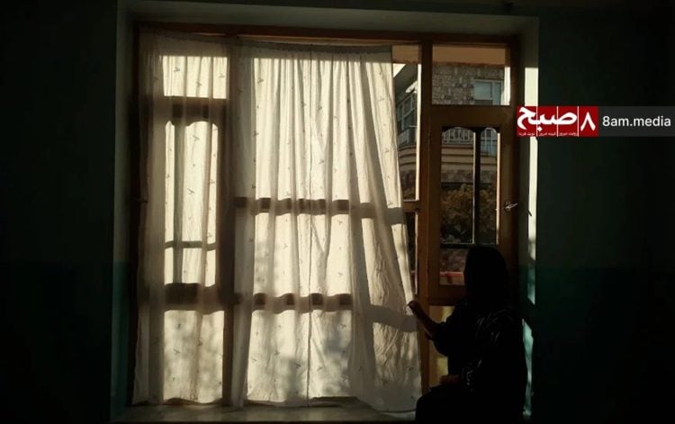 A silhouette of a woman stands before sunlit curtained windows in her modest home, embodying the quiet resilience of those bearing invisible burdens of loss and daily struggle. (Photo Credit: 8am.media)