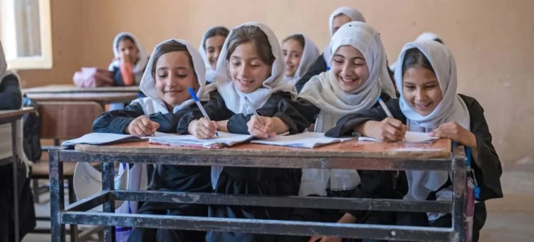 Young female students in hijabs smile while studying together at their classroom desks in Afghanistan, before sweeping education restrictions were imposed on girls and women. (Photo Credit: UNICEF)