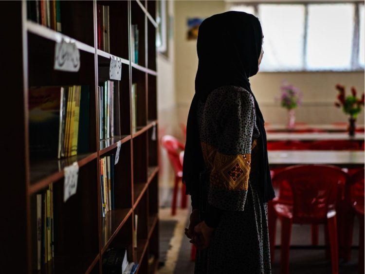 A young Afghan woman stands beside shelves of books in a library, symbolizing the enduring pursuit of education and knowledge despite restrictions on women's access to universities in Afghanistan. (Photo Credit: LA Times)
