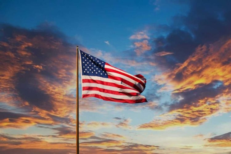 The American flag waves against a dramatic sunset sky, symbolizing U.S. global dominance and its role in shaping international order through military, political, and cultural influence across more than 700 bases worldwide. (Photo Credit: flagsusa)