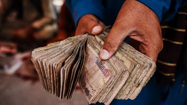 A Kabul resident holds a bundle of heavily worn banknotes, reflecting the growing cash crisis and the circulation of deteriorated currency in local markets. (Photo Credit: CA Times)