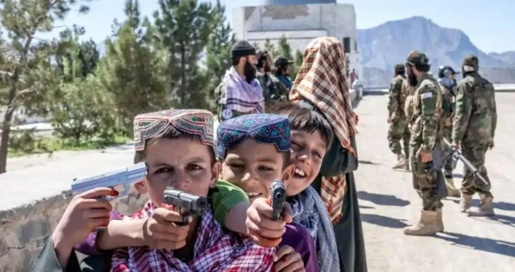 Young Afghan boys hold toy guns in Kabul as Taliban fighters stand in the background, illustrating growing concerns about the normalization of violence and militancy among children exposed to extremist propaganda. (Photo Credit: AP)