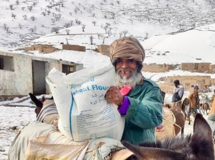 The image shows an elderly Afghan man smiling as he receives a sack of flour distributed by the United Nations. Photo Credit: WFP