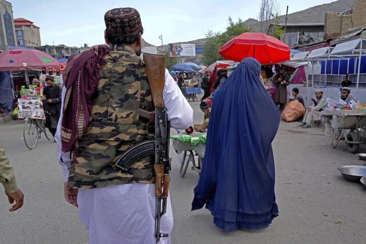 A Taliban fighter walks alongside a burqa-clad woman through a busy Afghan market, illustrating the enforced restrictions on women's movement and freedom under Taliban rule. (Photo Credit: AP)