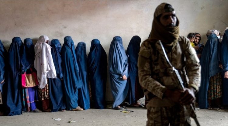 A Taliban fighter stands guard as Afghan women wait in line, illustrating the harsh restrictions stripping women of their basic rights and dignity across Afghanistan. (Photo Credit: AP)