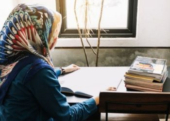 A young woman sits quietly at a desk, writing beside a stack of books and a sunlit window, carrying the quiet determination that defines countless Afghan women who fight for knowledge and dignity in silence. (Photo Credit: Pinterest)