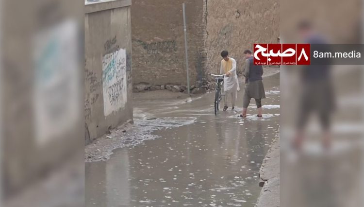 Two residents push a bicycle through a flooded street after rainfall in Kabul, highlighting water mismanagement challenges. (Photo Credit: 8am.media)
