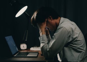A young professional sits exhausted at his desk late at night, head in hands, reflecting the growing toll of workplace stress and occupational depression among young workers. (Photo Credit: Getty Images)