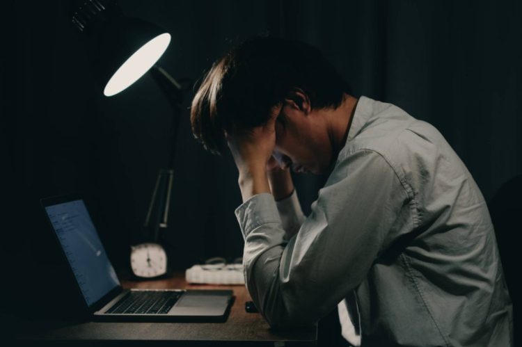 A young professional sits exhausted at his desk late at night, head in hands, reflecting the growing toll of workplace stress and occupational depression among young workers. (Photo Credit: Getty Images)