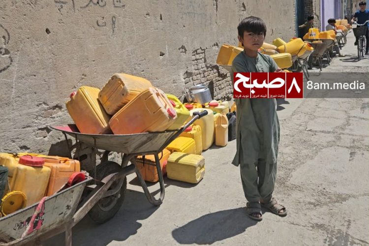 A boy stands beside wheelbarrows loaded with yellow jerry cans, lining up to collect water in Kabul. (Photo Credit: 8am.media)
