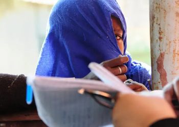 In this image, a young woman is shown with her face partially covered. Only one of her eyes is visible through the fabric, looking outward with a sense of concern, hesitation, or shyness. The overall atmosphere of the image is calm yet emotionally layered, reflecting quiet inner tension and unspoken feelings. (Photo credit: UNFPA)