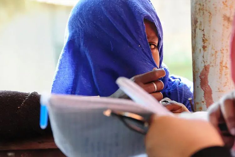 In this image, a young woman is shown with her face partially covered. Only one of her eyes is visible through the fabric, looking outward with a sense of concern, hesitation, or shyness. The overall atmosphere of the image is calm yet emotionally layered, reflecting quiet inner tension and unspoken feelings. (Photo credit: UNFPA)