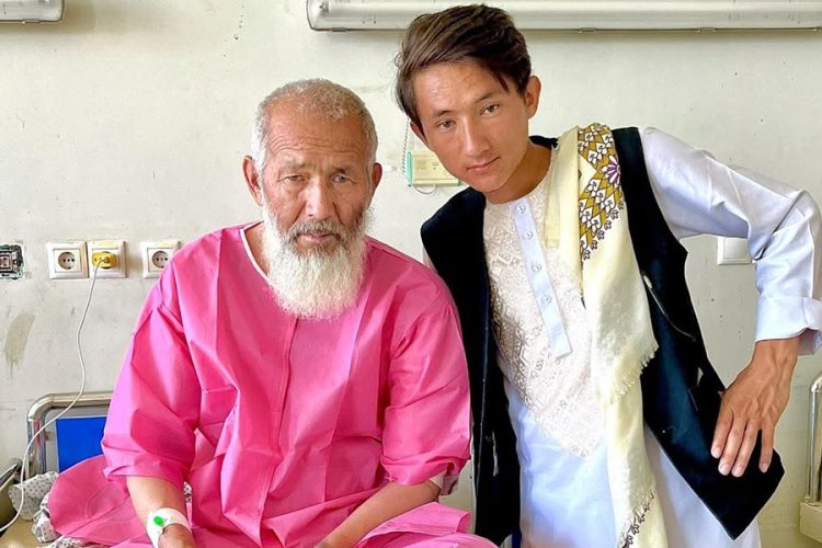 Esmat Khan Amiri sits with his father in a hospital ward amid allegations that he was denied surgery at a Kandahar hospital due to ethnic discrimination. (Photo credit: social media)