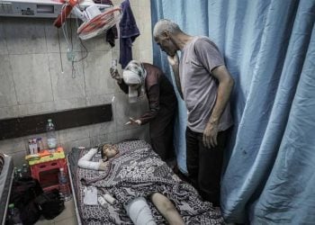A wounded child lies in a Gaza hospital bed, with bandaged limbs, as family members tend to them following an Israeli airstrike on a residential area in western Gaza. (Photo Credit: Al Jazeera)