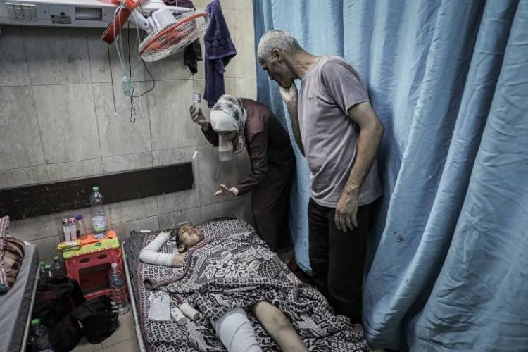 A wounded child lies in a Gaza hospital bed, with bandaged limbs, as family members tend to them following an Israeli airstrike on a residential area in western Gaza. (Photo Credit: Al Jazeera)