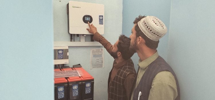 Two individuals inspecting and operating a solar power system and battery storage units supported by the United Nations Development Programme (UNDP), an initiative aimed at expanding access to sustainable energy and strengthening local power infrastructure. (Photo credit: UNDP)