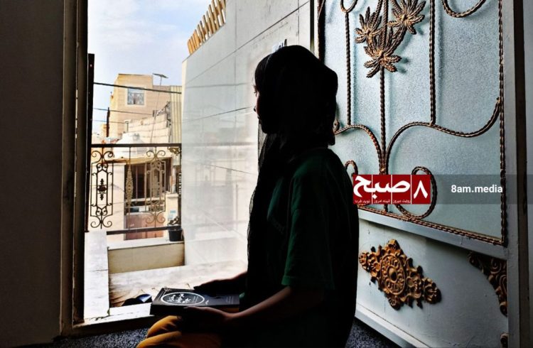 A schoolgirl sits at the entrance of her home holding books, reflecting on the continued closure of girls' schools in Afghanistan. (Photo credit: 8am.media)
