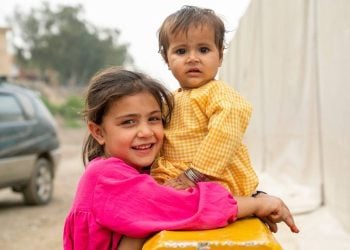 A young girl smiles as she holds her baby sister in her arms, sharing a quiet moment of affection and care (Photo: UNHCR)