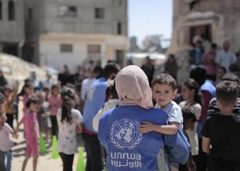 A UNRWA aid worker carries a child as families gather during the distribution of humanitarian assistance in the Gaza Strip. (Photo credit: UNRWA)