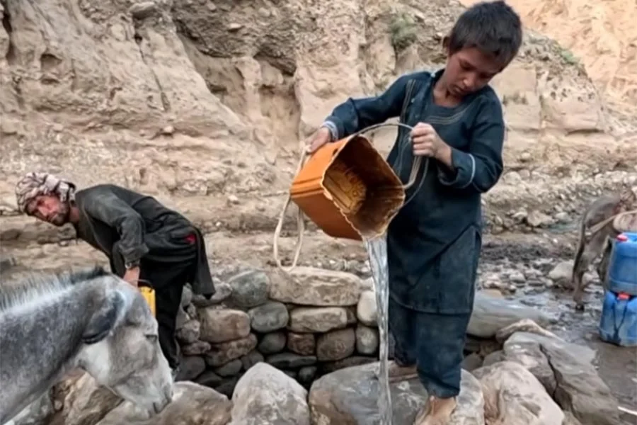 A boy pours water from a bucket at a village well as a man fills containers nearby with the help of a donkey. (Photo credit: Our Village)