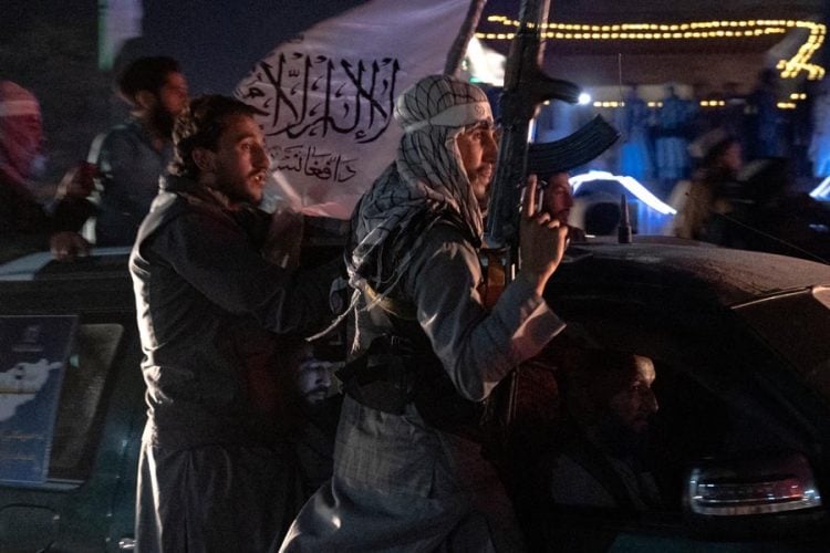 Armed Taliban fighters ride atop a vehicle at night, one carrying a rifle beneath a flag bearing Islamic script. (Photo credit: AFP)
