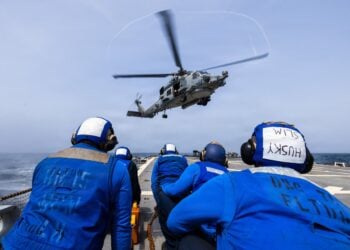 A military helicopter flies low over the flight deck of a naval vessel, while several crew members wearing bright blue shirts and protective flight deck helmets watch. (Photo: @CENTCOM/X)