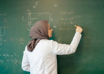 A female professor in a lab coat writes equations on a chalkboard, reflecting on a career cut short by the Taliban's ban on women's education. (Photo credit: Unsplash+)