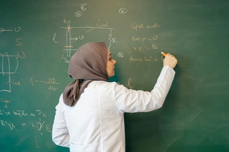 A female professor in a lab coat writes equations on a chalkboard, reflecting on a career cut short by the Taliban's ban on women's education. (Photo credit: Unsplash+)