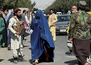 A Taliban fighter stands guard as a woman in a blue burqa crosses a street in Kabul. (Photo credit: Reuters)