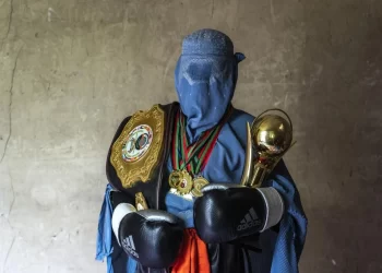 A woman in a burqa stands with medals, boxing gloves and a championship cup, highlighting women’s achievements in sports. (Photo credit: AP Photo/Ebrahim Noroozi)