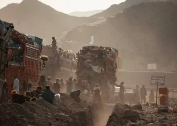 Afghan migrants gather near heavily loaded trucks along a dusty road in a mountainous area, carrying their belongings (Photo: IOM/Osman Azizi)