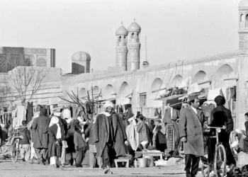 People walk through a crowded bazaar beside an arcaded wall and minarets in an Afghanistan city, in a historical black-and-white scene. (Photo credit: wikimedia)