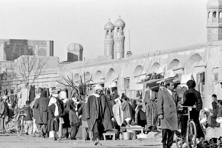 People walk through a crowded bazaar beside an arcaded wall and minarets in an Afghanistan city, in a historical black-and-white scene. (Photo credit: wikimedia)
