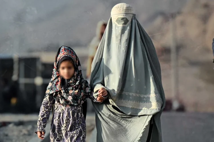 A woman in a burqa holds the hand of a young girl as they walk along a road in Afghanistan, where women heading households face poverty and social pressure. (Photo credit: AFP)